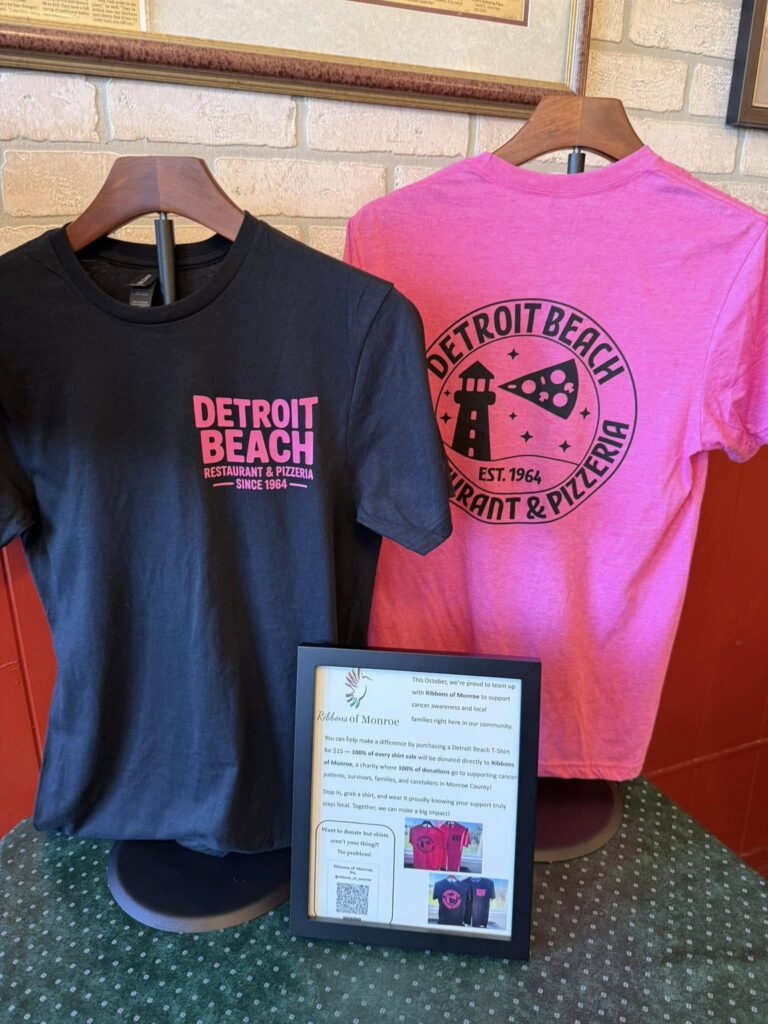 Display of black and pink t-shirts for the Detroit Beach Restaurant & Pizzeria fundraiser. A framed sign explains that 100% of shirt sales are donated to Ribbons of Monroe to support local cancer patients.