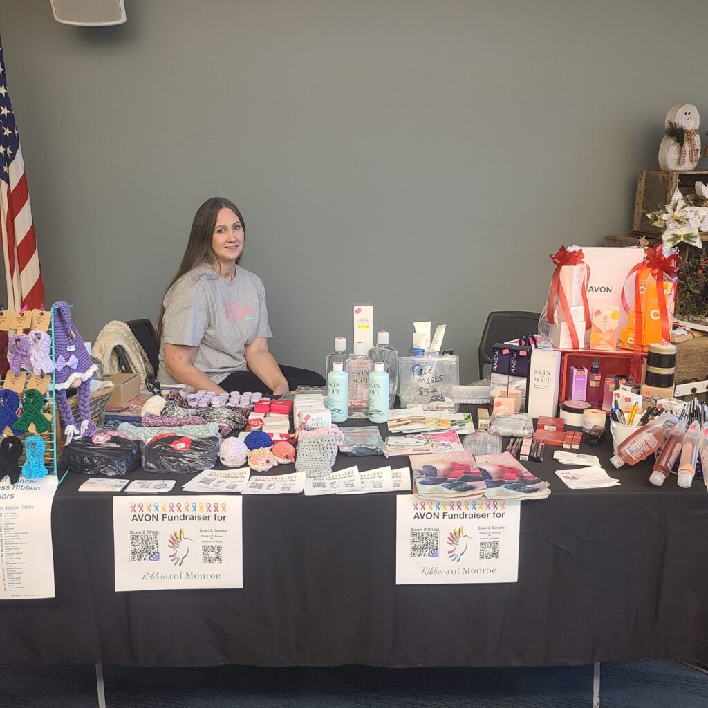 A volunteer sitting at a fundraising table for Ribbons of Monroe featuring Avon beauty products and handmade knitted cancer awareness ribbons. Signs display QR codes for direct donations.
